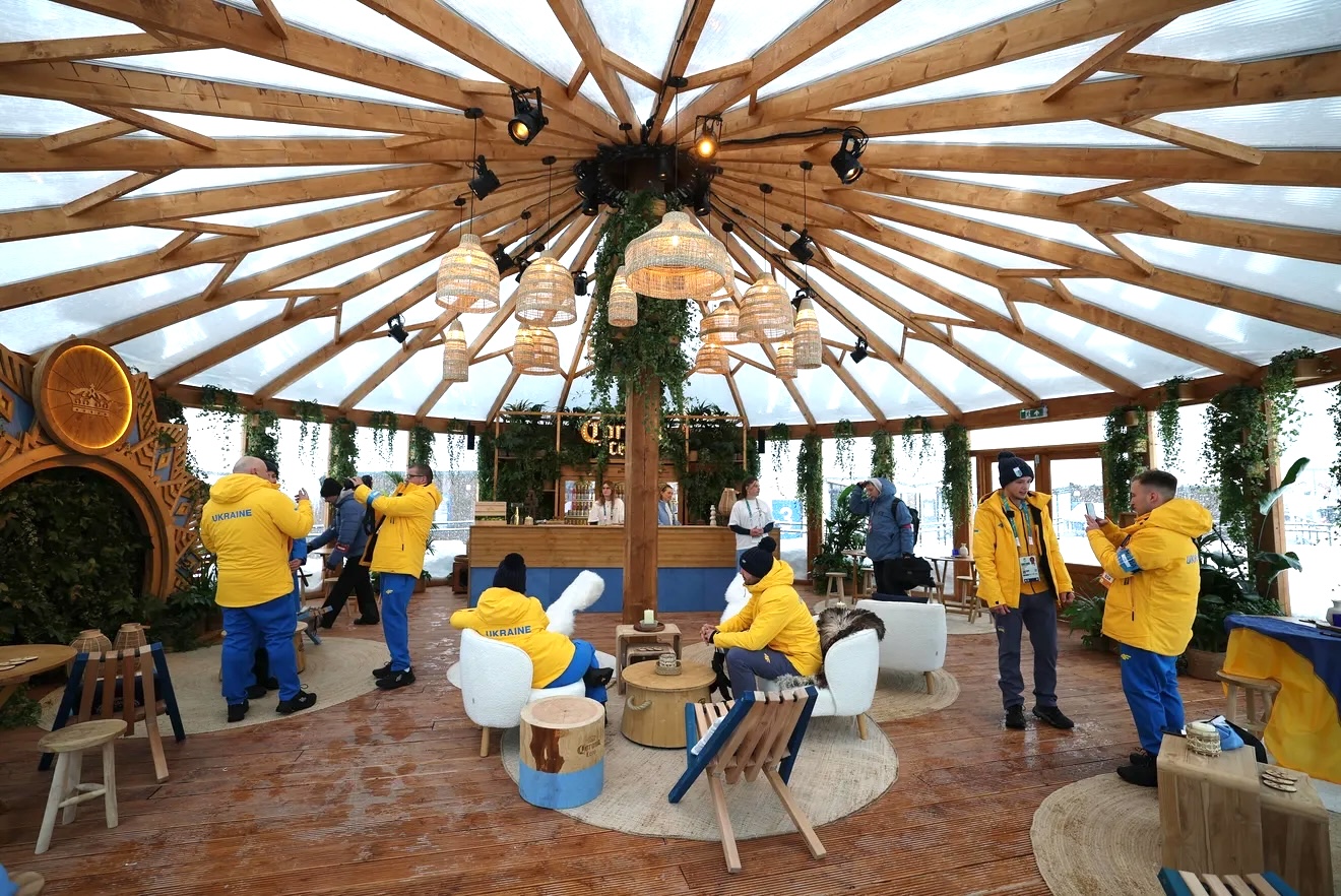 Team Ukraine athletes gather inside the Corona Cero Cantina during the Olympic Village Cortina Media Day on February 3, 2026 in Cortina, Italy. Dressed in bright yellow team jackets and blue pants, athletes relax and connect in the warm, wood-framed pavilion featuring woven pendant lights, greenery accents, and lounge seating arranged around a central column. Some athletes chat and check their phones while others pose for photos, creating a casual, behind-the-scenes moment amid the Olympic Village atmosphere.