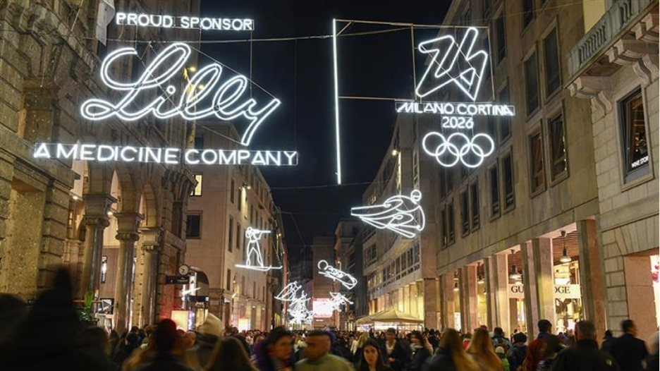 Nighttime view of a busy Milan shopping street illuminated with suspended light installations featuring winter sport figures, the Milano Cortina 2026 emblem and Olympic rings, and prominent “Lilly – A Medicine Company” sponsor signage glowing above the crowd.