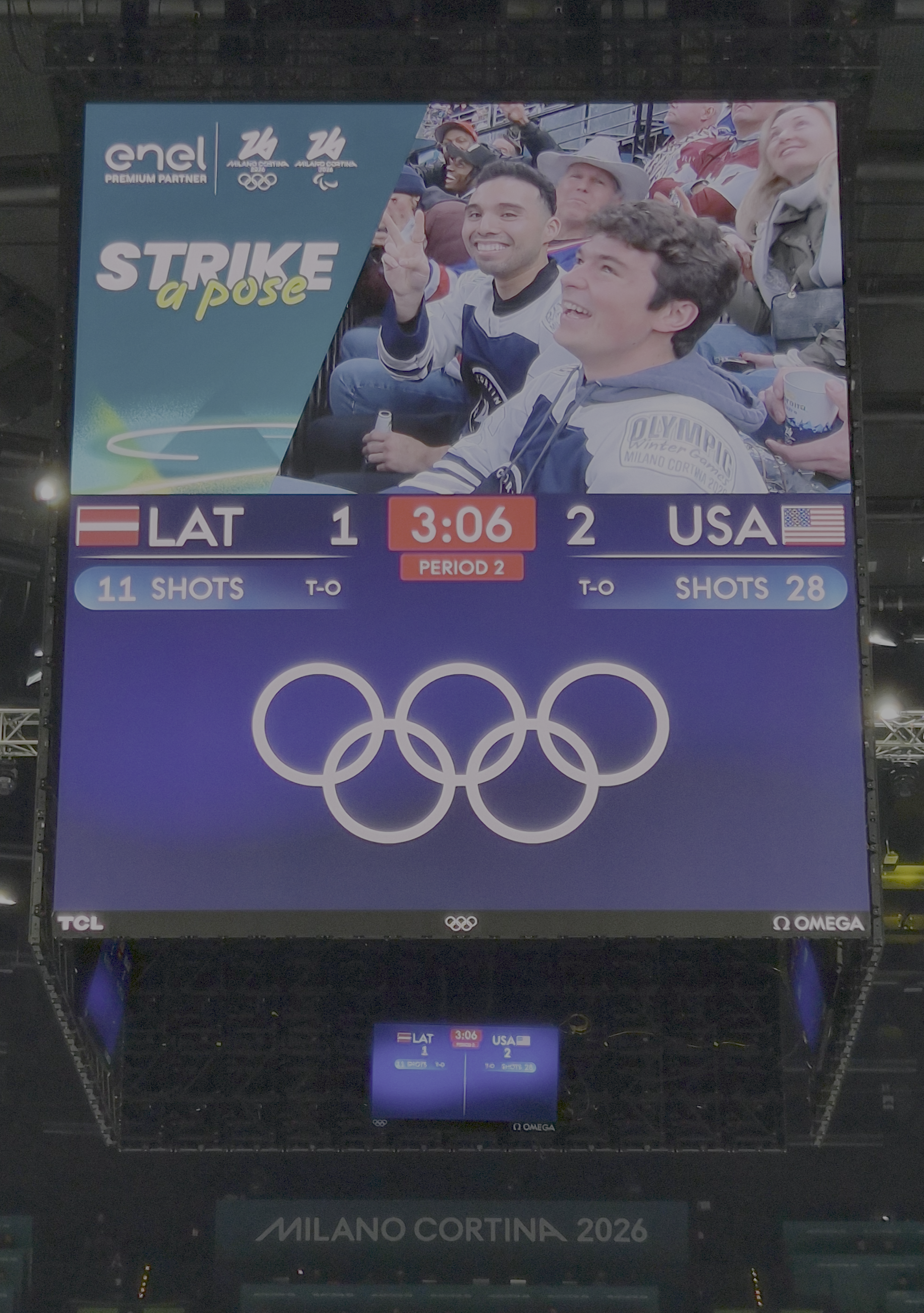Arena scoreboard at a Milano Cortina 2026 Olympic hockey game displaying Enel&rsquo;s &ldquo;Strike a Pose&rdquo; fan cam activation above the live score (USA 2, Latvia 1), with Olympic rings and sponsor logos visible inside the venue.